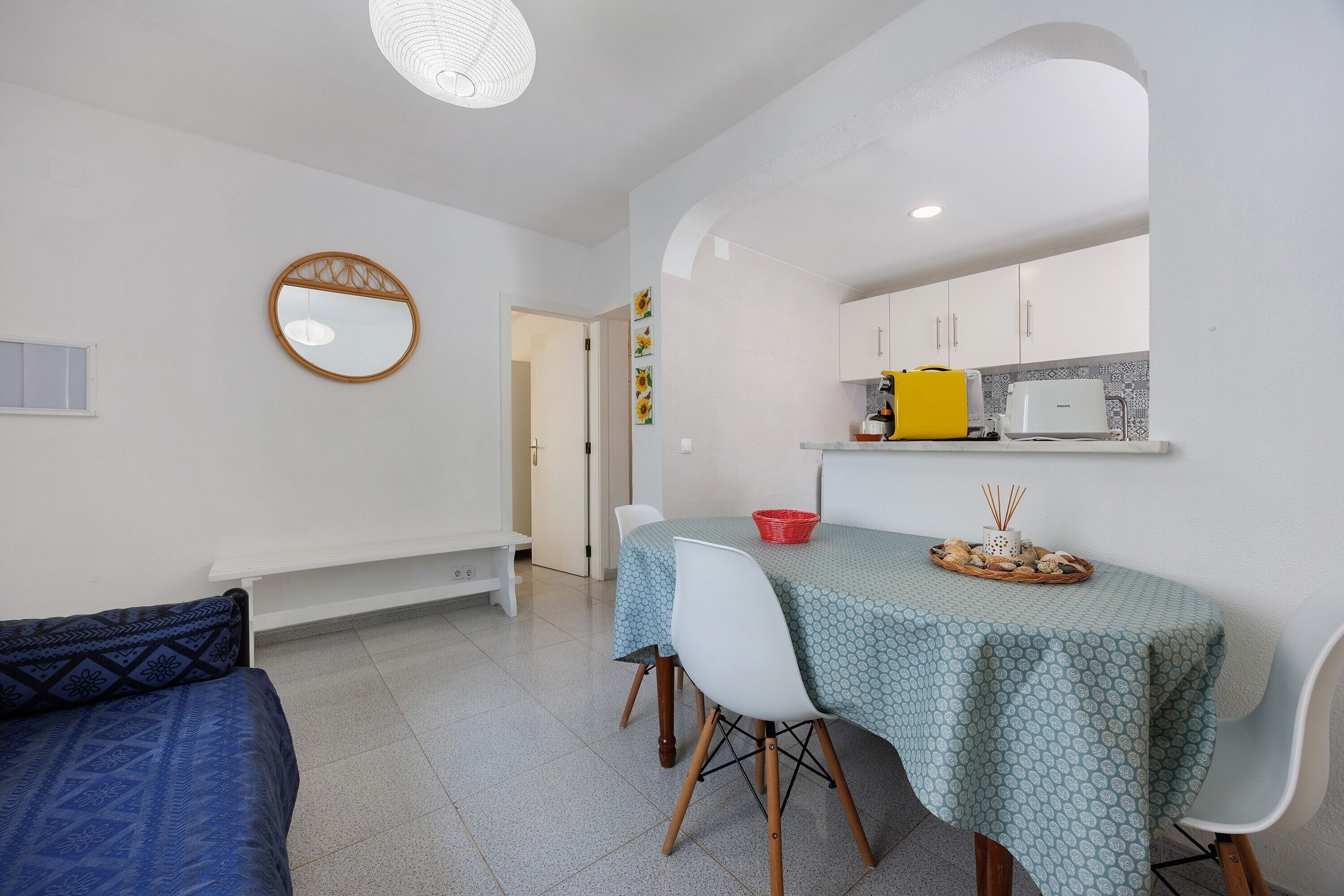 A compact living space with a dining table, chairs, a sofa, a kitchen area with cabinets, a mirror, and a ceiling light fixture.
