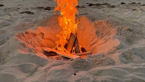 Ein Strand mit einer Feuerstelle im Sand, eine Person sitzt mit den Füßen im Sand, und im Hintergrund das Meer.
