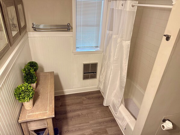 A bathroom with a wooden bench, potted plants, a window with blinds, a shower with a white curtain, and a towel rack.