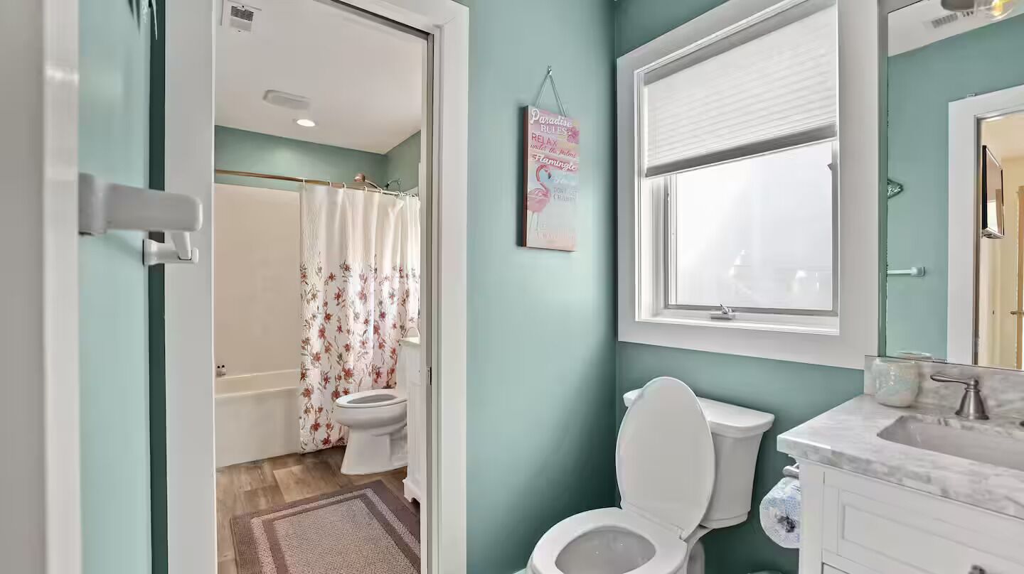 Ein Badezimmer mit Toilette, einer Dusche mit blumigen Vorhängen, einem Fenster und einem Waschbecken mit Marmorplatte.