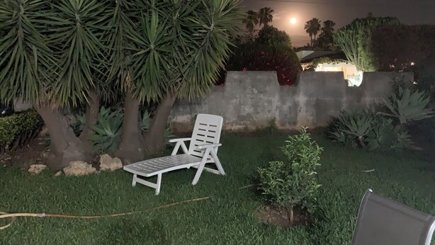 Un espacio exterior nocturno con una silla lounge blanca, palmeras y un muro de hormigón.