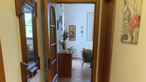 Un pasillo con puerta de madera, un espejo y suelo de baldosas que conduce a un comedor con una silla de mimbre y una mesa.