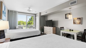 A hotel room with two beds, a TV, a desk, and a balcony with a view of trees.