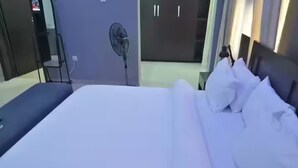 Un llit ben fet amb roba de llit blanca, una taula de nit amb una làmpada i un ventilador a la paret.