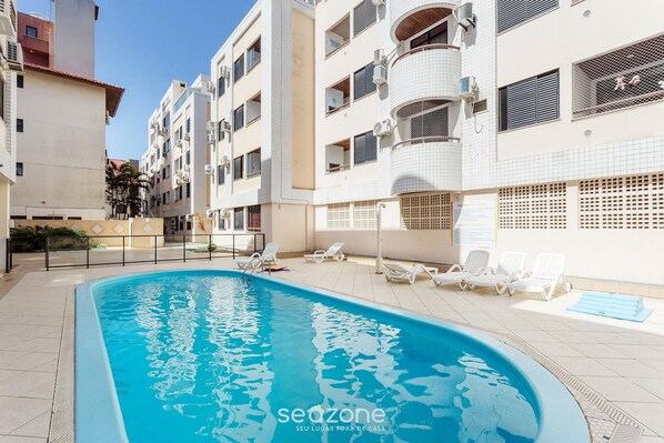 Uma piscina com espreguiçadeiras e um deck revestido em azulejos, cercada por edifícios de vários andares com varandas e aparelhos de ar condicionado.
