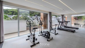A modern gym with stationary bikes and treadmills, large windows with a city view, and a clean, minimalist design. - Apartment With Barbecue in Itacorubi Aft0210 (Florianópolis)