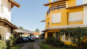 A residential area with a yellow building, a covered patio, parked cars, and a pathway. - House in Condo w/ Pool, 300m From the Beach Ali024 (Florianópolis)