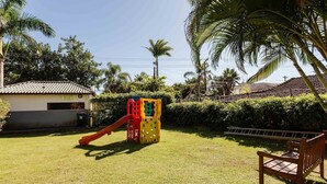 A playground with a slide and climbing structure, surrounded by grass and palm trees. - House in Condo w/ Pool, 300m From the Beach Ali024 (Florianópolis)