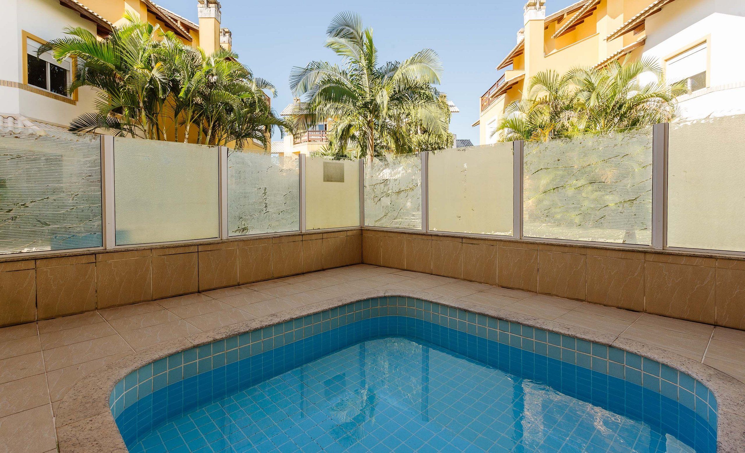 Una piscina con bordes de azulejos, rodeada de un piso y paredes cubiertos de baldosas beige, con una partición de vidrio y palmeras al fondo.