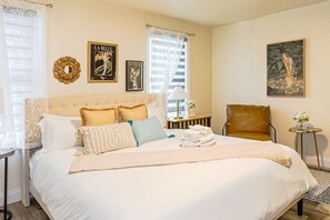 Une chambre à coucher élégamment aménagée, avec un grand lit, une tête de lit, une table de chevet avec une lampe, un fauteuil et des œuvres d’art accrochées aux murs.