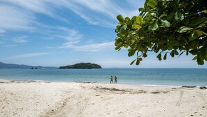 Una playa con arena, una pequeña isla a lo lejos y dos personas caminando por la orilla.