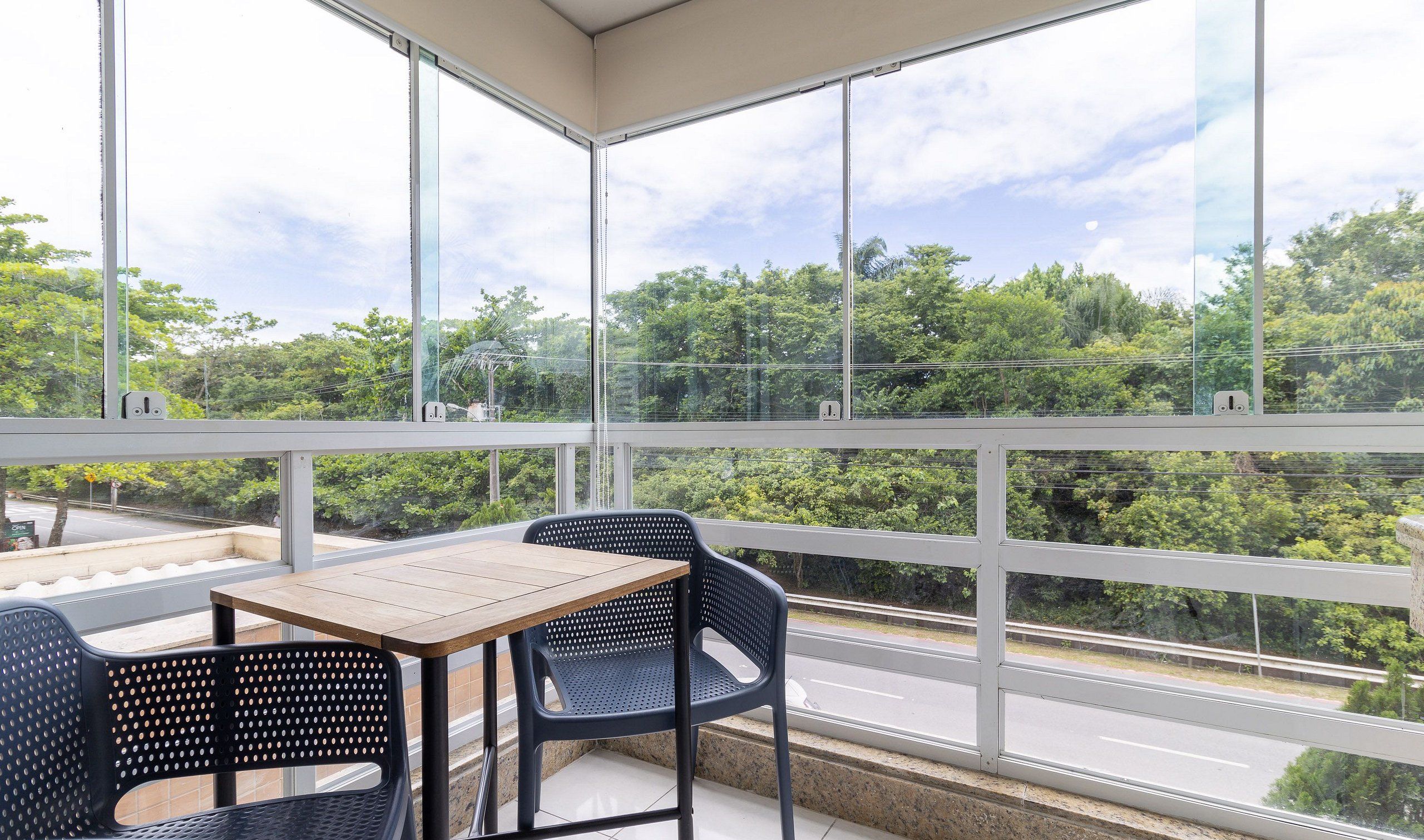 A balcony with a table and chairs, large windows offering a view of a road and greenery.