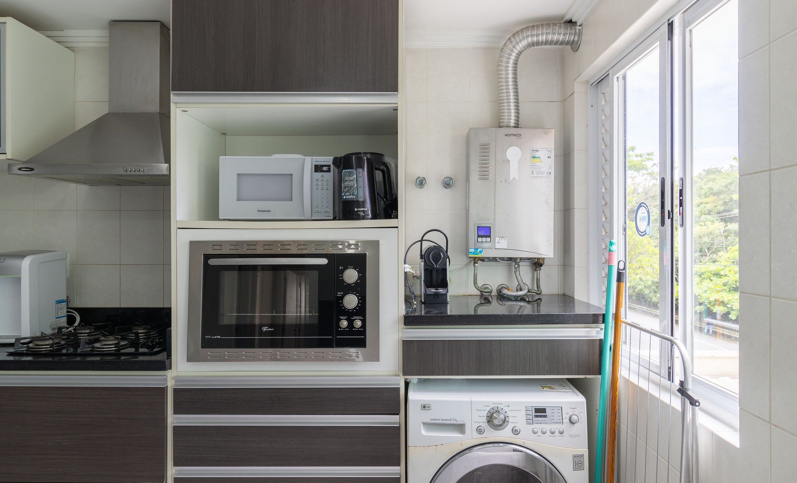 A compact kitchen with a built-in microwave, oven, and washing machine, featuring a window with a view of the outdoors.