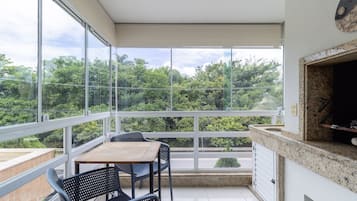 A balcony with a wooden table and chairs, a kitchen area with a countertop, and large windows offering a view of greenery.