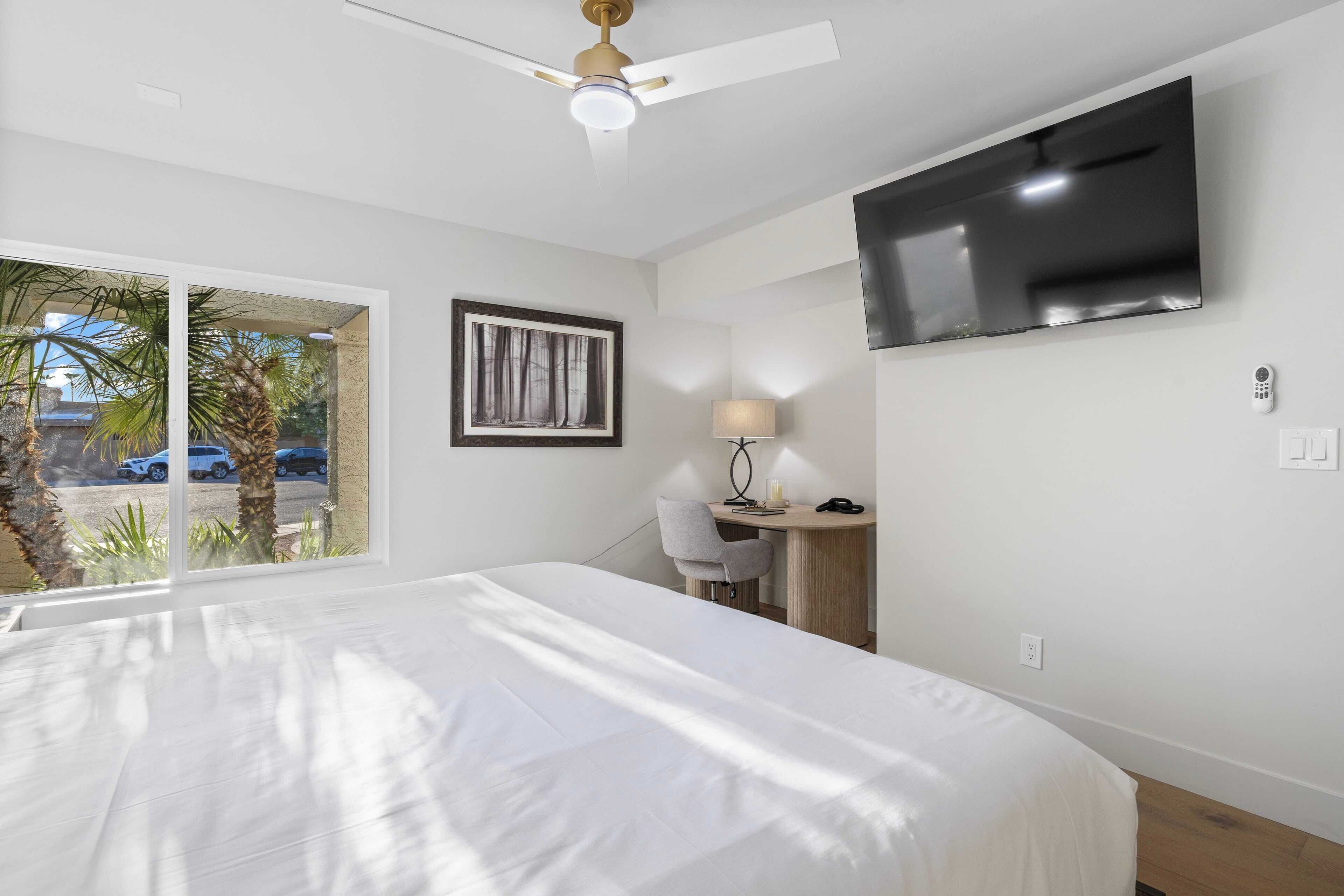 A hotel room with a bed, a desk, a chair, a TV, and a window with a view of palm trees and cars.