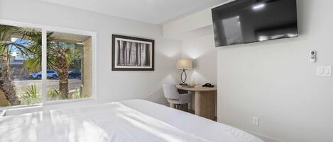 A hotel room with a bed, a desk, a chair, a TV, and a window with a view of palm trees and cars.