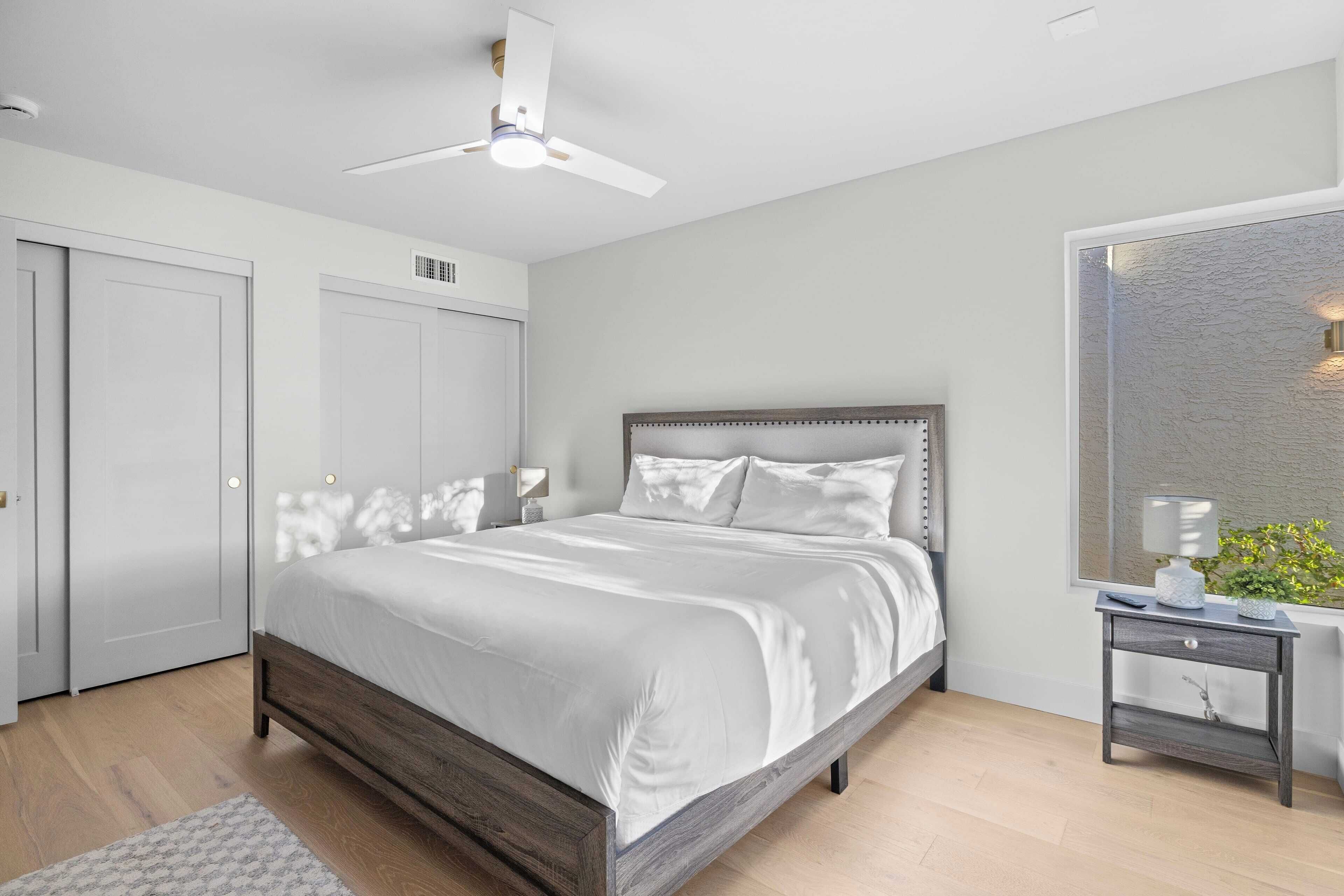 A modern bedroom with a large bed, a ceiling fan, a nightstand, and a window with a view.