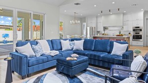 A modern living room with a large blue sectional sofa, a small ottoman, and a coffee table. There is a kitchen area in the background with white cabinets and stainless steel appliances. - El Dorado by Avantstay Luxe 4BR w/ Pool, Putting Green, Fireplace Near Kierland (Scottsdale)