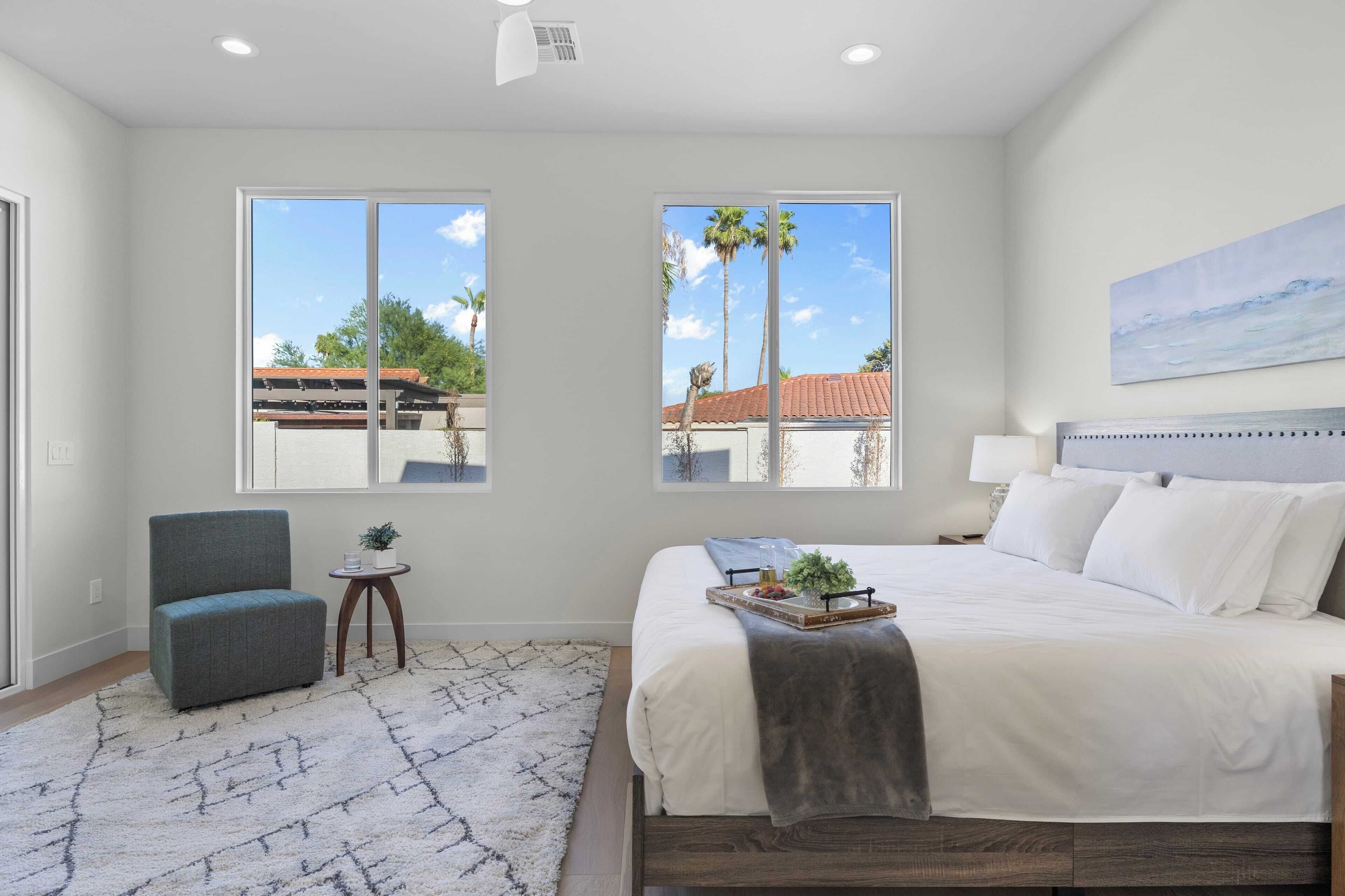 A bedroom with a large bed, two windows, a chair, a small table, and a painting on the wall.