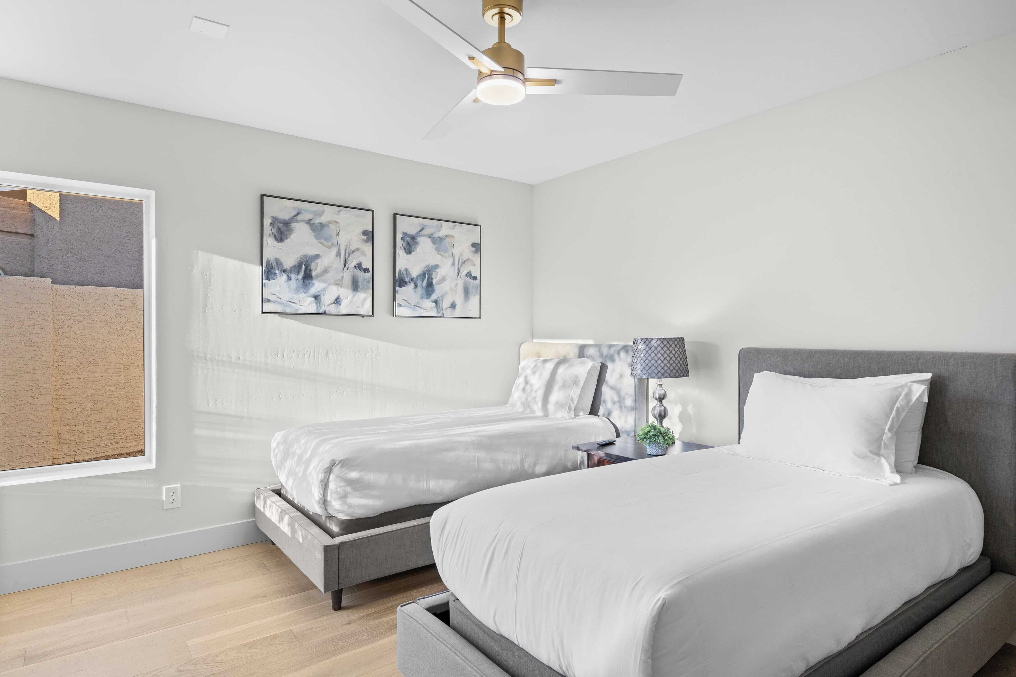 A bedroom with two beds, a ceiling fan, a window with a view, and abstract wall art.