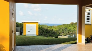 A yellow building with a white door, a stone patio, and a view of a green landscape and ocean.