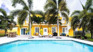 A swimming pool with a yellow building in the background, palm trees, and outdoor seating.