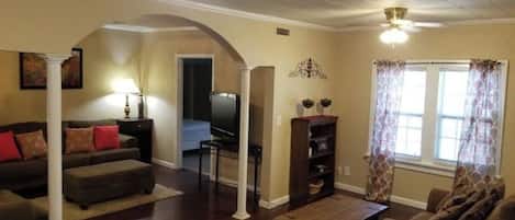 Un salon comprenant un canapé, un pouf, une table basse et un meuble pour la télévision. Il y a une fenêtre avec des rideaux et un ventilateur de plafond.