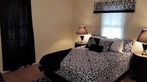 Een slaapkamer met een bed, twee bedlampen, een raam met gordijnen met een patroon, en een zwart-wit beddekleed met een patroon.