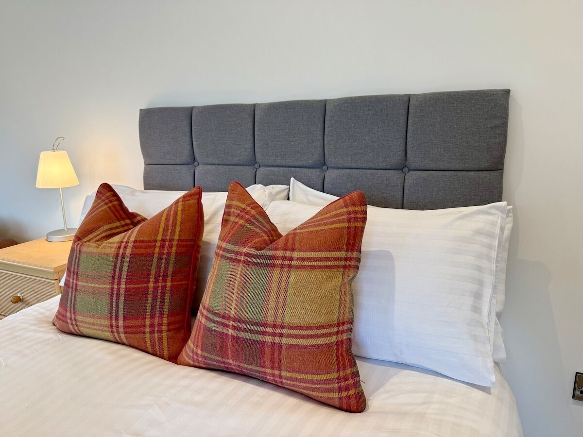 A bed with a grey upholstered headboard, white bedding, and plaid pillows.