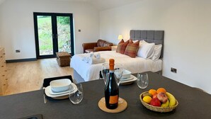 A modern bedroom with a bed, bedside table with wine and fruit, and a view of the outdoors. - Retreat In The Hills - The Old Post Office (Malvern)