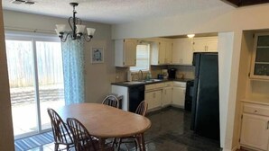 A compact kitchen with a dining table and chairs, a refrigerator, and a window with blinds. - Crestwood Bungalow (Stillwater)