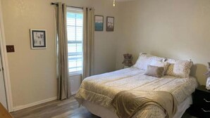 A bedroom with a large bed, a window with curtains, and a wooden floor. - Crestwood Bungalow (Stillwater)