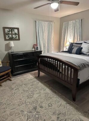 A bedroom with a bed, a dresser, a nightstand, a ceiling fan, and patterned carpet.