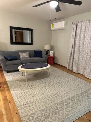 A living room with a grey sofa, a round coffee table, a patterned rug, a ceiling fan, and a window with curtains.