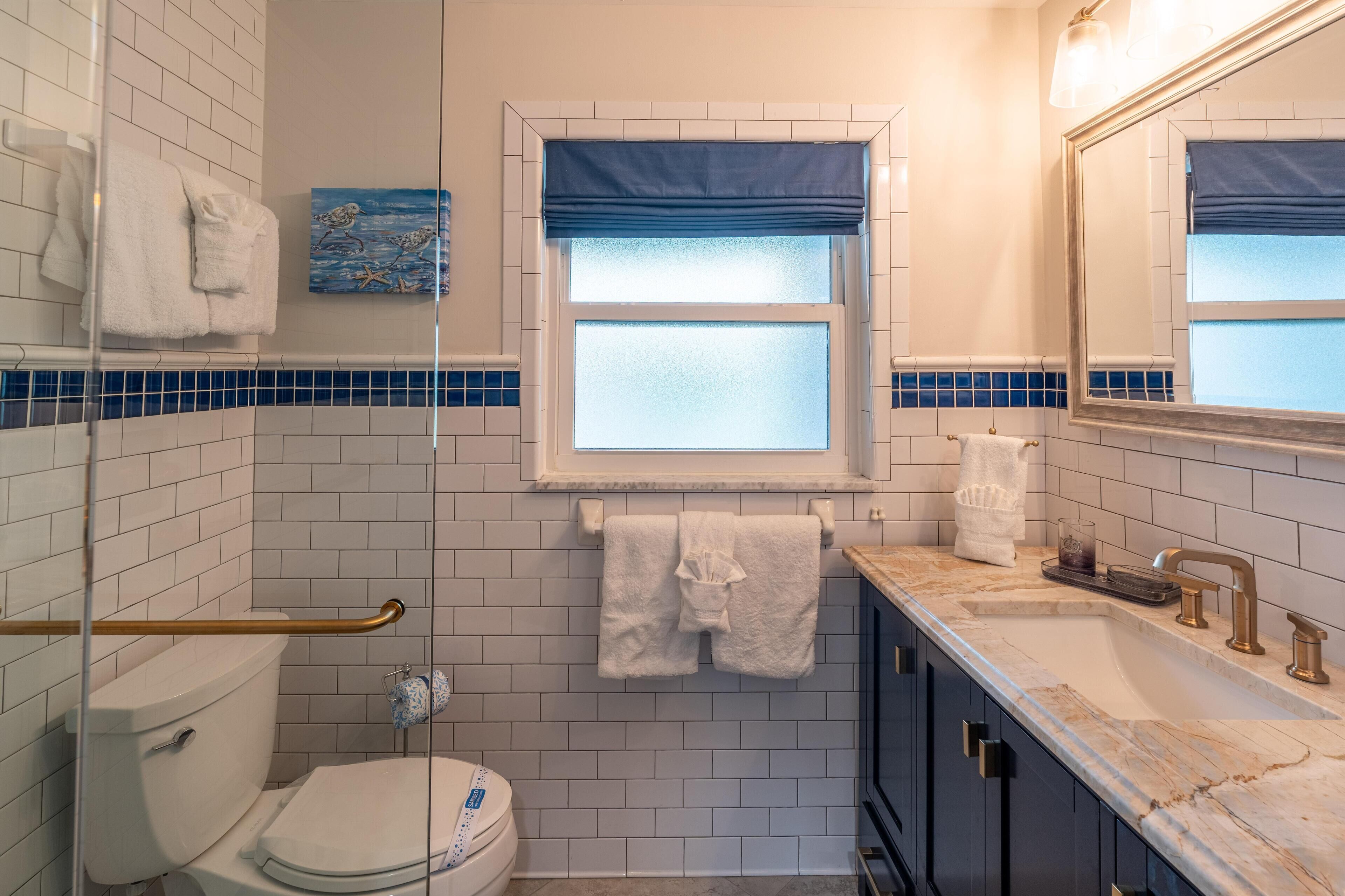 A bathroom with white tiles, a window with blue blinds, a white toilet, a sink with a mirror, and a towel rack.