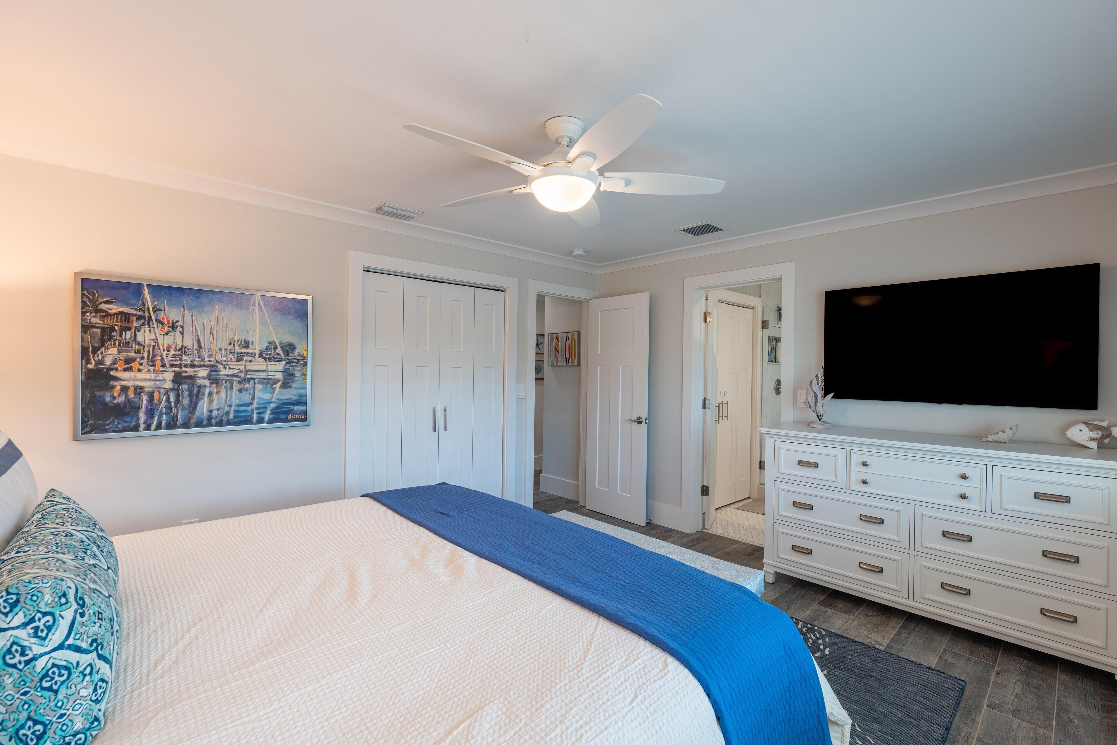 A bedroom with a bed, a ceiling fan, a TV, and a painting on the wall.