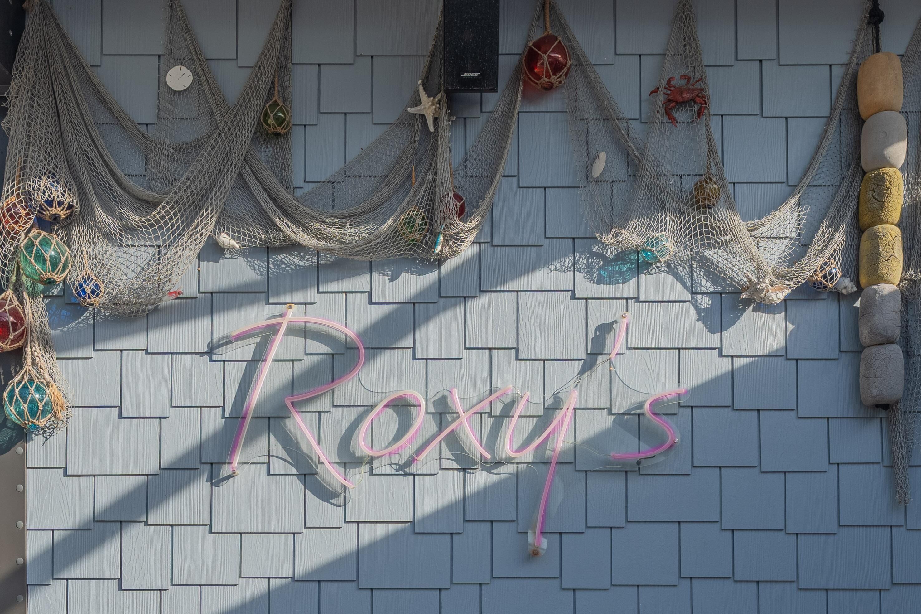 A wall with a neon sign spelling "Roxy's" and various fishing nets and weights hanging above it.