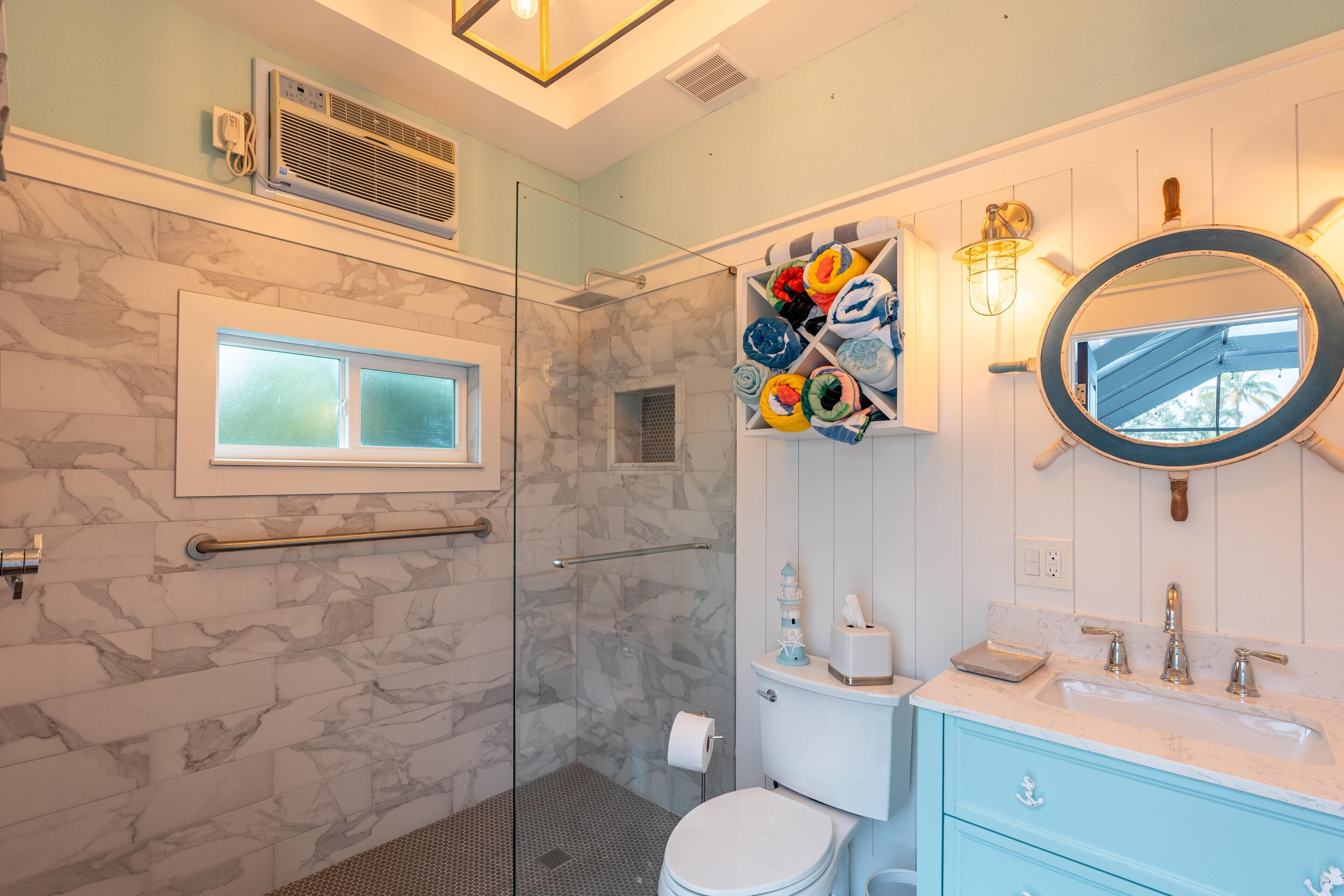 A bathroom with a glass shower enclosure, a toilet, a sink with a mirror, and a towel rack with colorful towels.