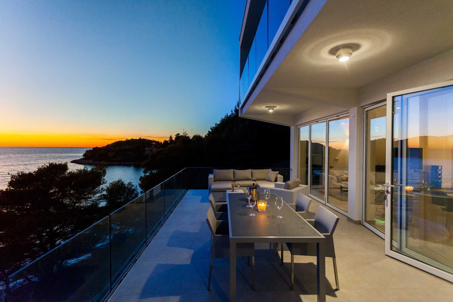 A modern outdoor dining area with a view of the sunset over the sea, a glass balustrade, and a sofa with cushions.