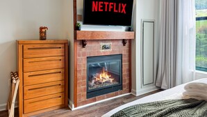 A room with a fireplace, a television mounted above it displaying the Netflix logo, a wooden cabinet, and a bed with a green blanket. - Heated Pool Hot Tubs Sauna Gym Mtn Front Appalachian @ Mtn Creek #428 (Vernon)
