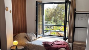 Une chambre avec deux lits, une table de chevet avec une lampe, une fenêtre avec des rideaux et une vue sur les arbres et les fleurs à l’extérieur.