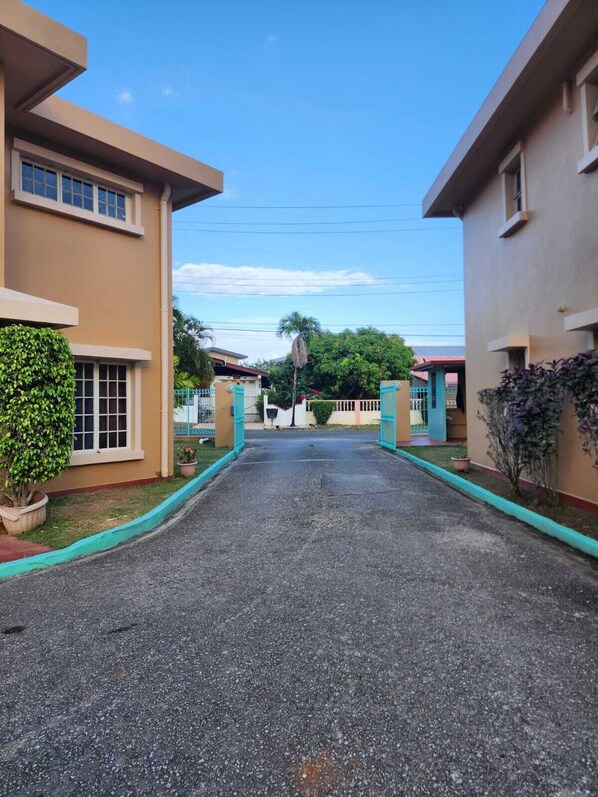 Una calle residencial con casas de dos pisos, cielo despejado y una vía central que conduce a una puerta.