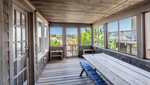 Une véranda couverte avec des bancs en bois et une table, de grandes fenêtres donnant sur une clôture et de la verdure.