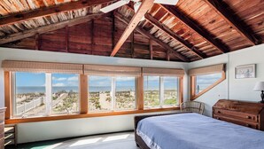 Une chambre avec une grande fenêtre, des poutres apparentes en bois au plafond, un lit, une commode et une chaise.