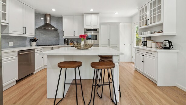 Una cocina moderna con armarios blancos, una isla central con taburetes, una campana extractora de acero inoxidable y un salpicadero.