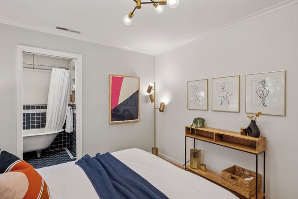 Un dormitorio con una cama, una mesita de noche con lámpara, estante en la pared y un baño visible a través de una puerta abierta.