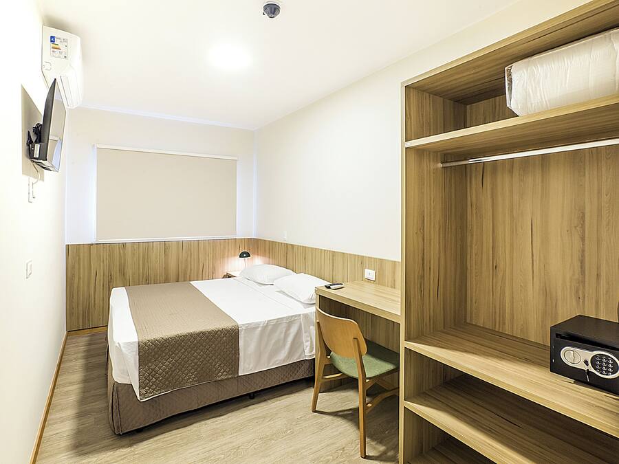A compact hotel room with a bed, a desk, a chair, and a shelf with a radio.