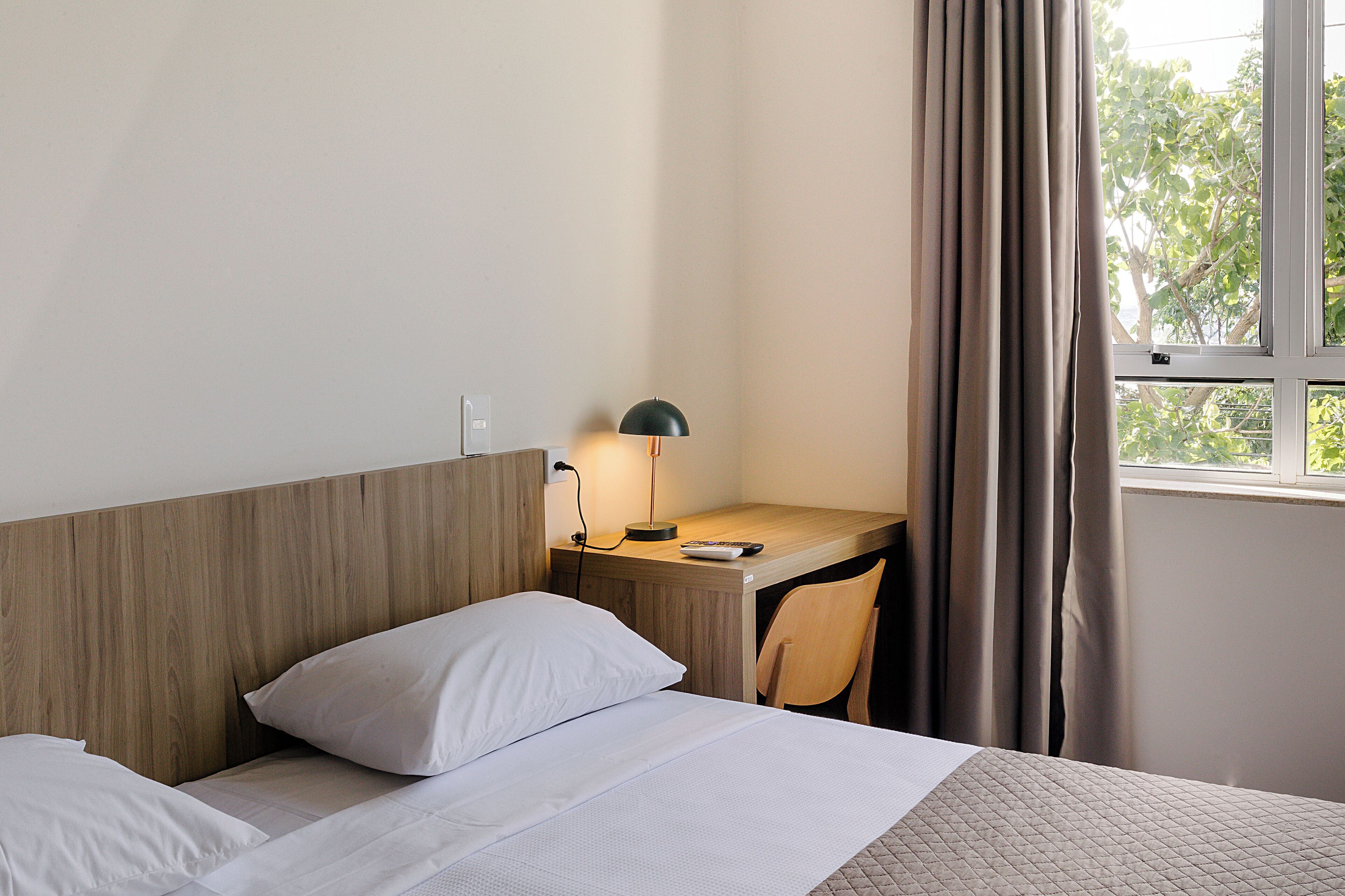 A hotel room with a wooden headboard, a desk with a lamp, a chair, and a window with curtains.