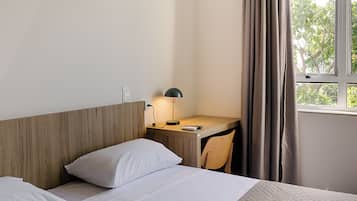 A hotel room with a wooden headboard, a desk with a lamp, a chair, and a window with curtains.