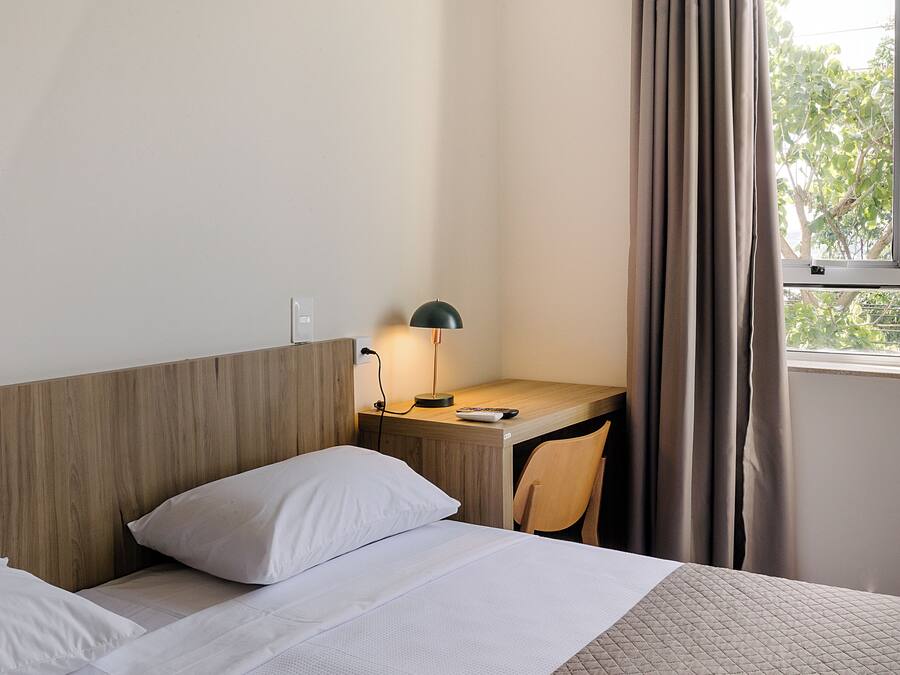 A hotel room with a wooden headboard, a desk with a lamp, a chair, and a window with curtains.